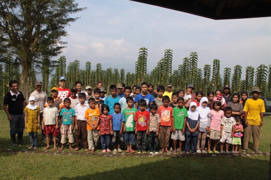 A group portrait of the children sponsored by the estate alongside their parents, the plantation workers, in front of a row of young teak with mountains in the background.